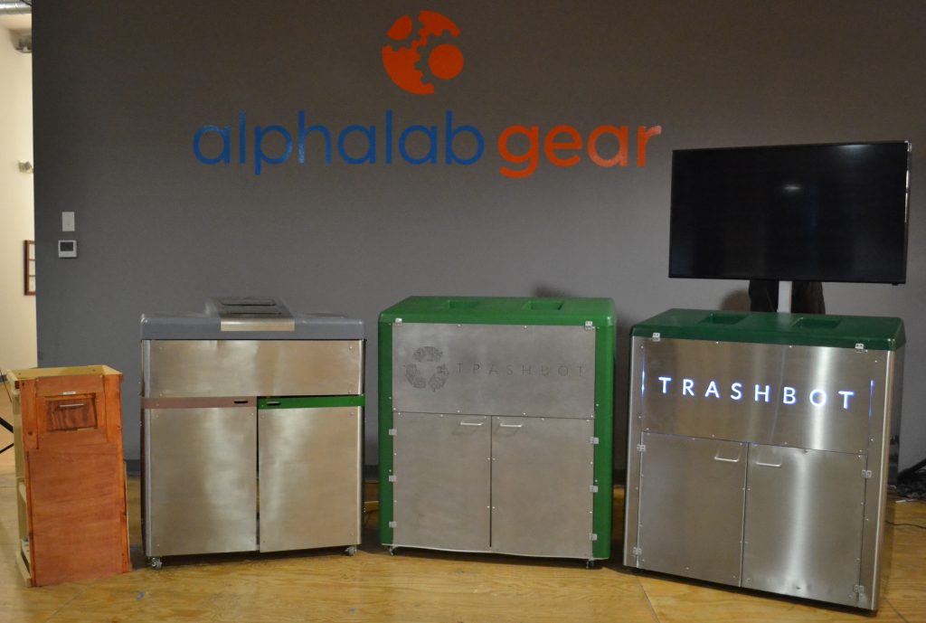 A picture of all the different designs of TrashBot. Starting with the original wooding prototype, then a prototype hacked out of an outdoor grill, then the front/back design with recycling logo, and finally the current version with side-by-side trash deposit holes.